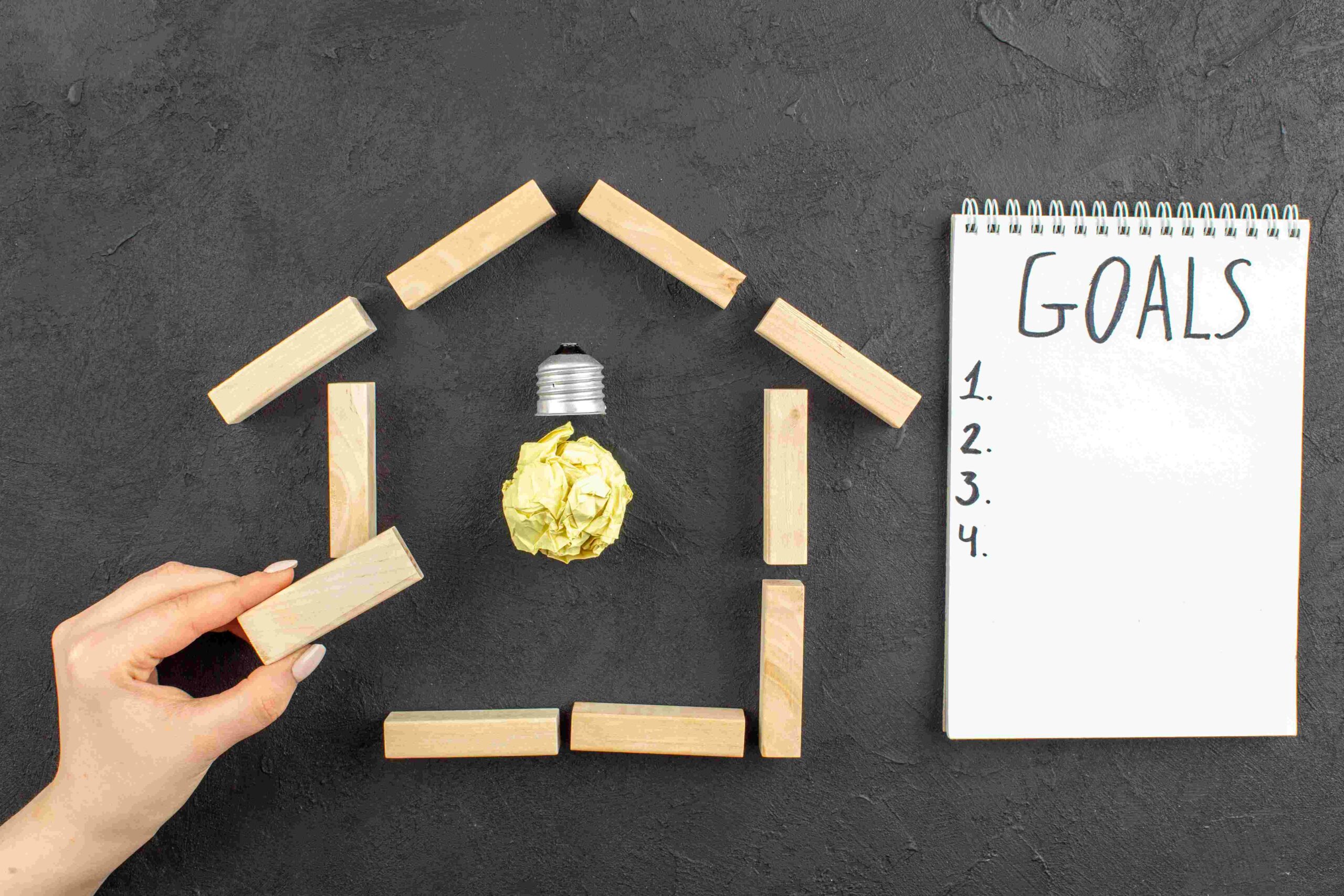 compressed_top-view-idealight-bulb-house-shaped-wood-blocks-goals-written-notepad-wood-block-female-hand-black